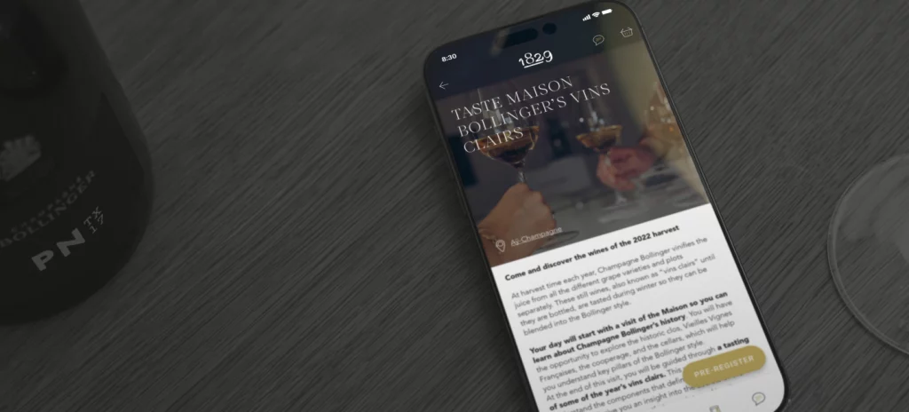 Un smartphone affichant une application avec un article intitulé "TASTE MAISON BOLLINGER'S VINS CHAIS" en contexte d'une dégustation de vins, posé sur une table en bois sombre, à côté d'une bouteille et un verre à vin.