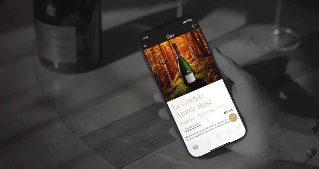 Une main tient un smartphone affichant une bouteille de vin avec texte: «La Grande Année Rosé Magnum - Millésime 2015, 600 €». En arrière-plan, une table avec une bouteille et un verre.