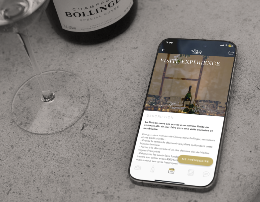 Un smartphone affiche des informations sur une visite d'expérience, posé sur une table en béton à côté d'un verre et d'une bouteille de Champagne Bollinger. Texte à l'écran: VISITE EXPERIENCE DESCRIPTION La Maison ouvre ses portes à un nombre limité de visiteurs afin de faire vivre une visite exclusive et mémorable. Plongez dans l’univers de Champagne Bollinger, ses secrets de fabrication, et ses savoir-faire. - Visites privées menées par des guides fondateurs et histo... Meilleur Accueil de France dans la vallée depuis 2001.